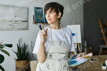 Mature woman artist reflects in her vibrant art studio while holding paintbrushes 58 Mature woman artist reflects in her vibrant art studio while holding paintbrushes