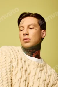Young man with distinctive tattoos poses thoughtfully against a vibrant yellow backdrop