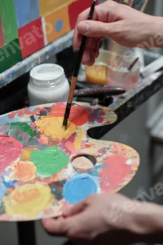 Hand of young professional painter mixing colors on palette in front of easel